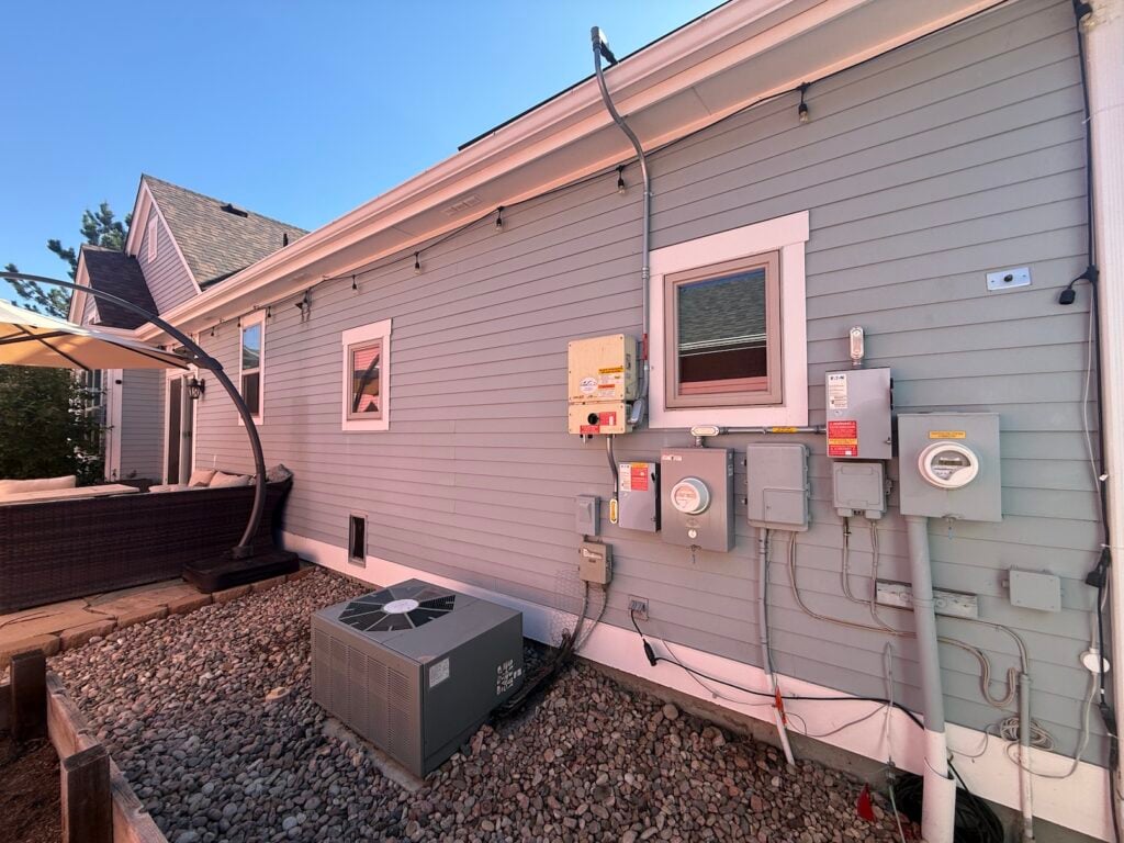 Exterior side of Robert Germain's home before heat pump install, showing the old outdoor AC condenser unit sitting on gravel next to the electrical meters, solar disconnect, and utility boxes.