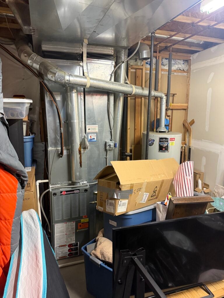 Basement mechanical room before install, showing the existing Rheem gas furnace and Bradford White gas water heater with sheet-metal ductwork above.