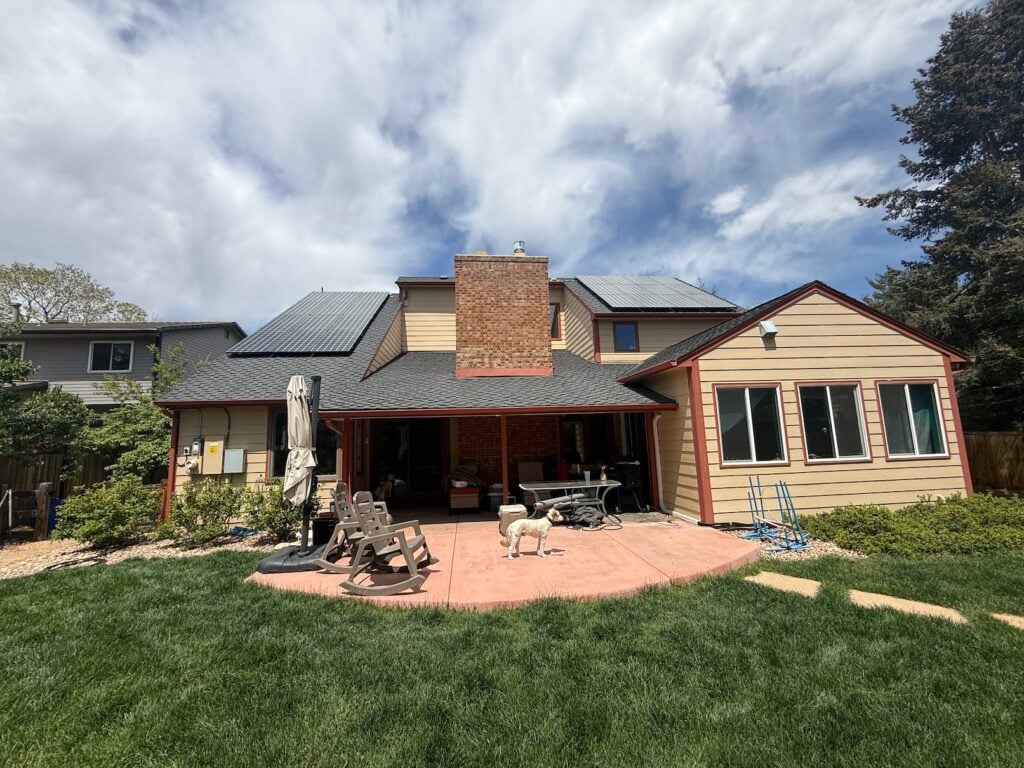 Rear exterior of David's Lakewood home before the heat pump install, a two-story tan house with rooftop solar panels, a large brick chimney, and a covered patio.
