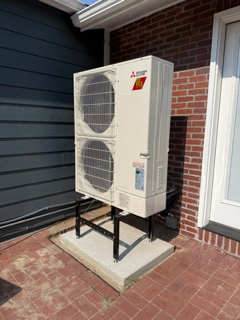New Mitsubishi Electric heat pump condenser mounted on a raised stand and concrete pad beside a brick home in Denver after installation