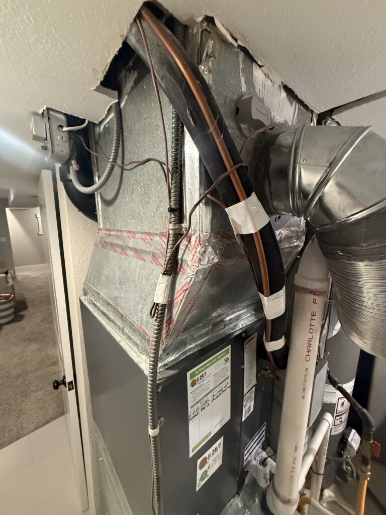 Close-up of an existing furnace with refrigerant lines and sheet-metal ductwork in a Denver home before heat pump installation