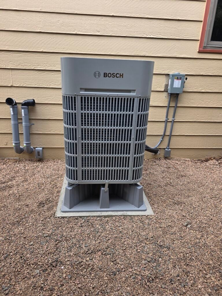 New Bosch outdoor heat pump unit installed at David's home, mounted on a gray composite pad on gravel next to the electrical disconnect and PVC intake/exhaust piping.